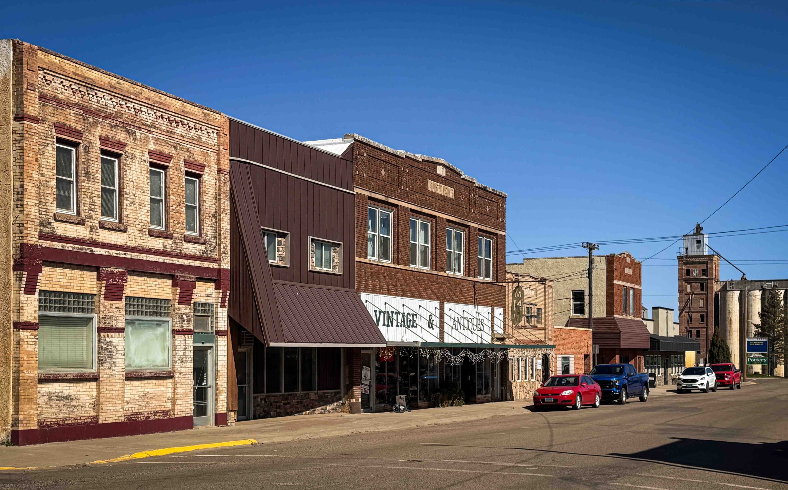 A small-town main street with vintage brick buildings, a shop with a striped awning, parked cars, and a clear blue sky. A taller historic building is visible in the background. Beach, SD