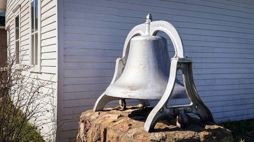 A large, silver metal bell sits atop a rough stone base outside a white wooden building with horizontal siding and a window. Shrubs are visible nearby on a sunny day. Beach, SD