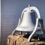 A large, silver metal bell sits atop a rough stone base outside a white wooden building with horizontal siding and a window. Shrubs are visible nearby on a sunny day. Beach, SD