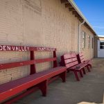 Red wooden benches labeled “GOLDEN VALLEY COUNTY MUSEUM” are lined up against a beige brick wall outside a building, with a white door in the background under a clear blue sky. Beach, SD