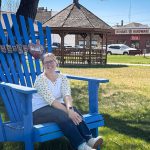 A smiling person with glasses sits on an oversized blue wooden chair in a grassy park area, with a gazebo, trees, and a hardware store visible in the background. A “Beach” sign hangs on the chair. Beach, SD