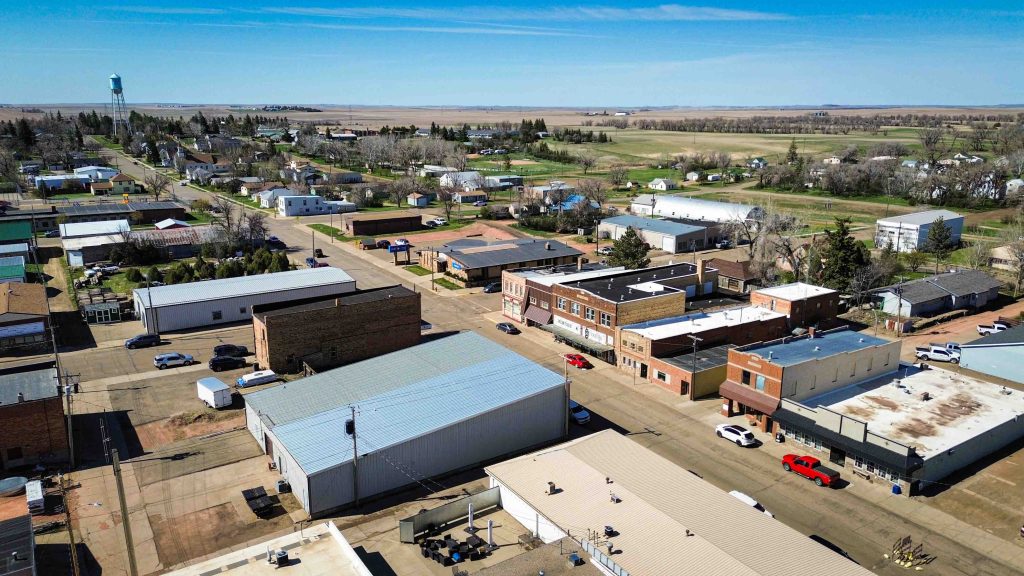 Aerial view of a small rural town with low-rise buildings, cars parked along the streets, and fields in the distance under a clear blue sky. Beach, SD