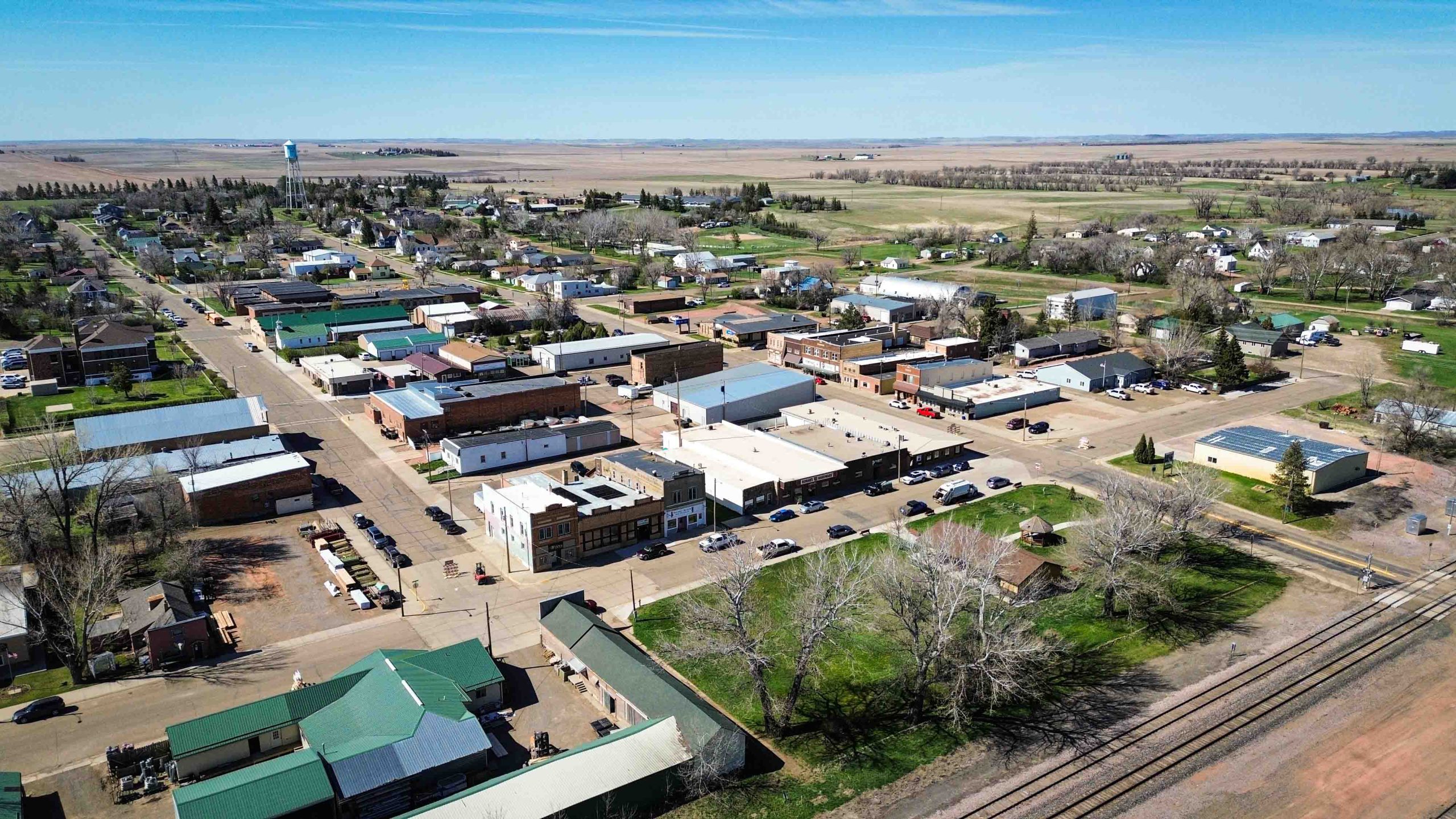 Aerial view of a small rural town with low-rise buildings, residential houses, streets with parked cars, green lawns, scattered trees, and open farmland in the background under a clear blue sky. Beach, SD