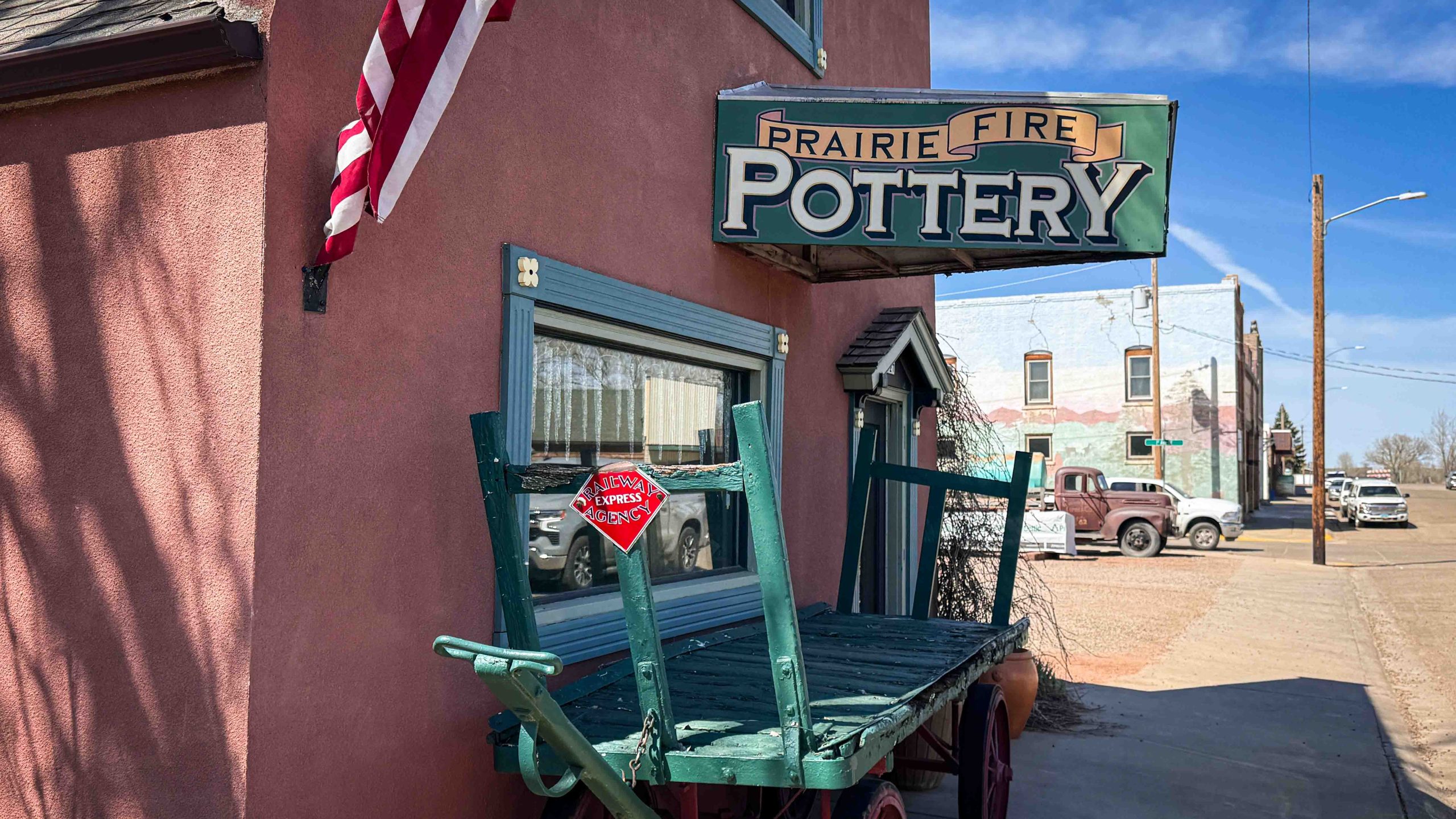 A green wooden cart with a red Look! Please Do Not Climb sign is in front of a Prairie Fire Pottery shop with an American flag and vintage cars visible on a sunny street. Beach, SD