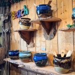 Colorful ceramic pots with wooden spoons are displayed on rustic wooden shelves and a wall. A colorful beach sign and other pottery items are arranged in a cozy, wood-paneled shop. Beach, SD