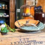 A handcrafted ceramic plate and a small vase with a white flower sit on a rustic wooden crate inside a cozy shop with shelves of pottery and wooden decor in the background. Beach, SD