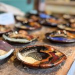 A close-up of colorful handmade ceramic plates displayed on a rustic wooden table, with blurred plates and pottery in the background. Beach, SD