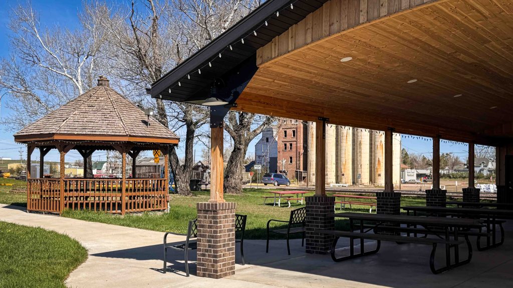 A park with a wooden gazebo, picnic tables, and a covered pavilion. Tall silos and a brick building are visible in the background, along with leafless trees and a clear blue sky. Beach, SD