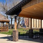 A park with a wooden gazebo, picnic tables, and a covered pavilion. Tall silos and a brick building are visible in the background, along with leafless trees and a clear blue sky. Beach, SD