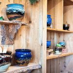 Handmade ceramic pots and bowls with earthy tones and blue glaze are displayed on rustic wooden shelves, with some wooden utensils placed in a pot. The display has a cozy, natural, artisanal feel. Beach, SD