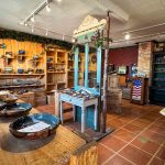 Artisanal pottery shop interior with wooden shelves and tables displaying various ceramic bowls and pottery pieces. The space has warm lighting, terra-cotta tile floors, and rustic decor with vintage and handmade touches. Beach, SD