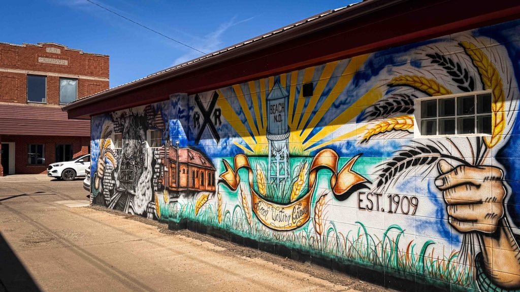 A colorful mural on a brick wall in Beach ND depicts wheat, a train, a grain elevator labeled York, and the text EST. 1909, celebrating local agricultural history under a bright blue sky.