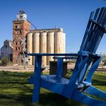 A large blue Adirondack chair sits on grass in the foreground, with grain silos, an old industrial building, and a railroad crossing sign visible in the background under a clear blue sky. Beach, SD