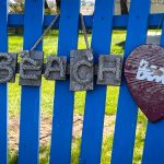A blue wooden fence displays hanging decor: four square plaques spelling BEACH with rope and shells, and a red heart-shaped sign with partially faded white text. Green grass and tree trunks are visible behind the fence. Beach, SD